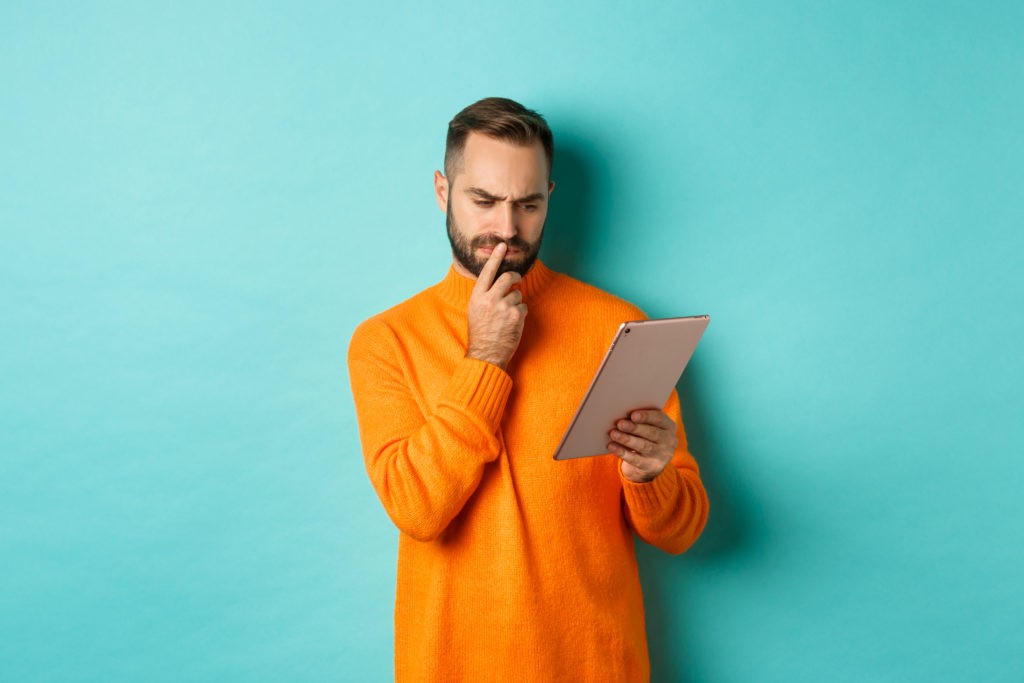 Um homem em dúvida com tablet na mão, um suéter laranja, barba e de costas para um fundo azul  Pré-vendas