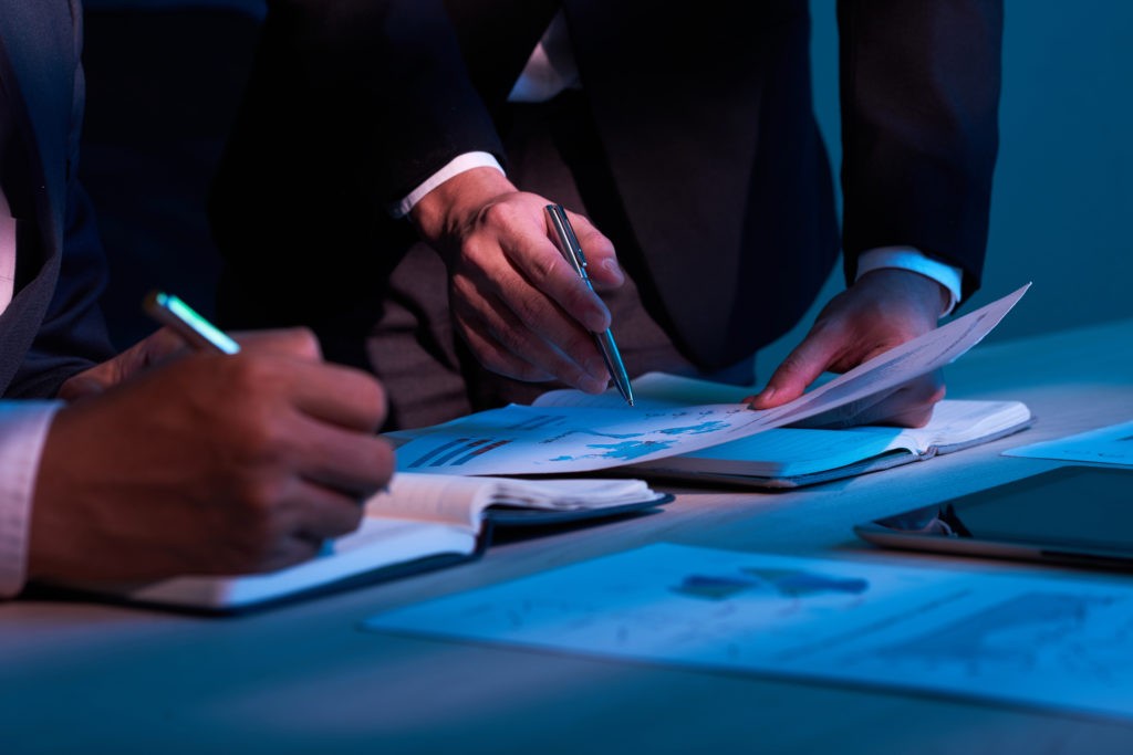 Imagem de dois executivos em frente a uma mesa em ambiente de iluminação azul. 

O foco está nas mãos dos dois que seguram cada uma caneta. Um com um caderno e outro com um mapa. 

Estratégias de vendas para varejo