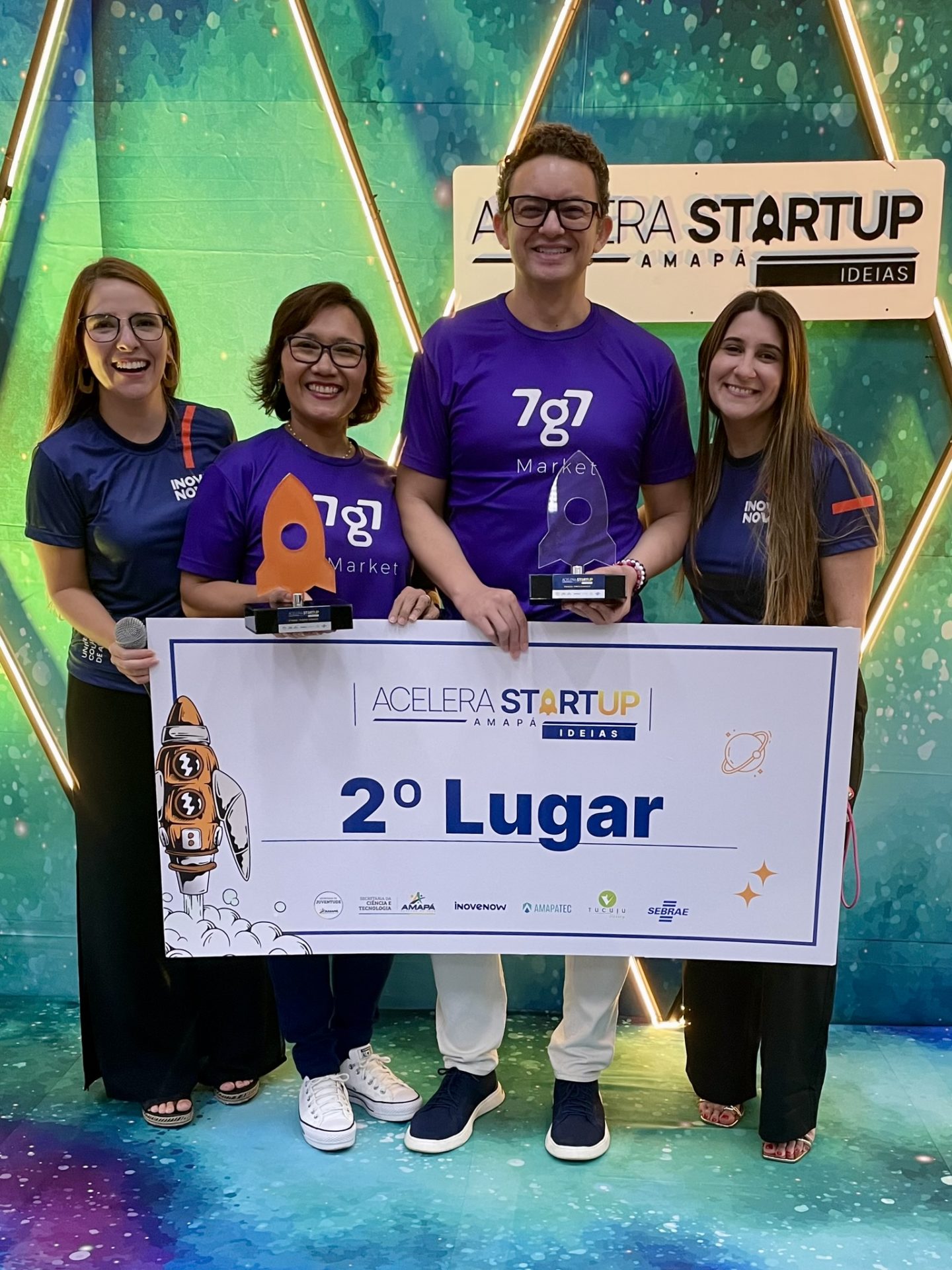 Quatro participantes exibem troféus e placa de segundo lugar no Demo Day 2023, diante do painel do Acelera Startup.