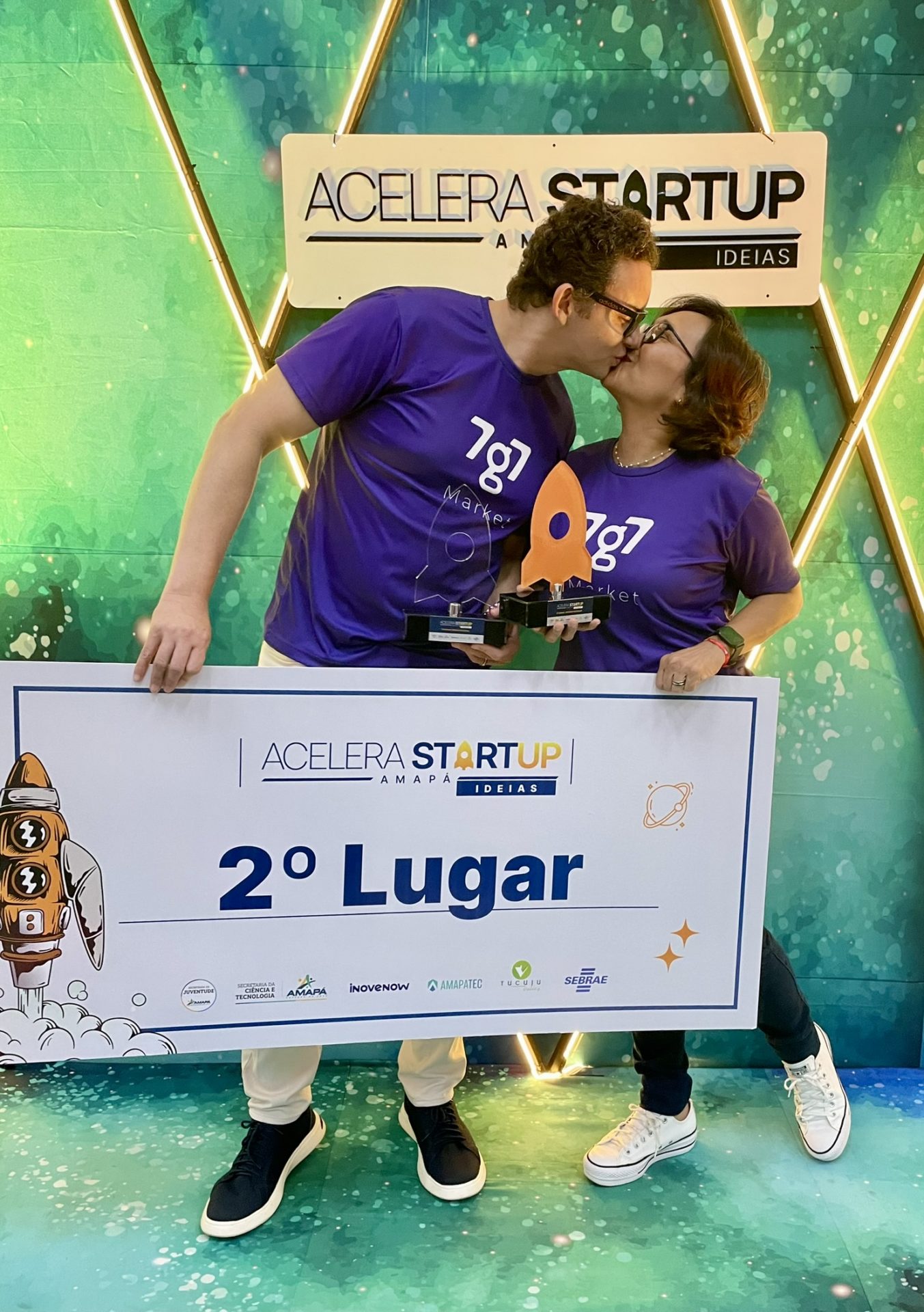 Maikon Richardson e Tania Assunção se beijam segurando troféus atrás da placa de segundo lugar do Acelera Startup, em 2023.