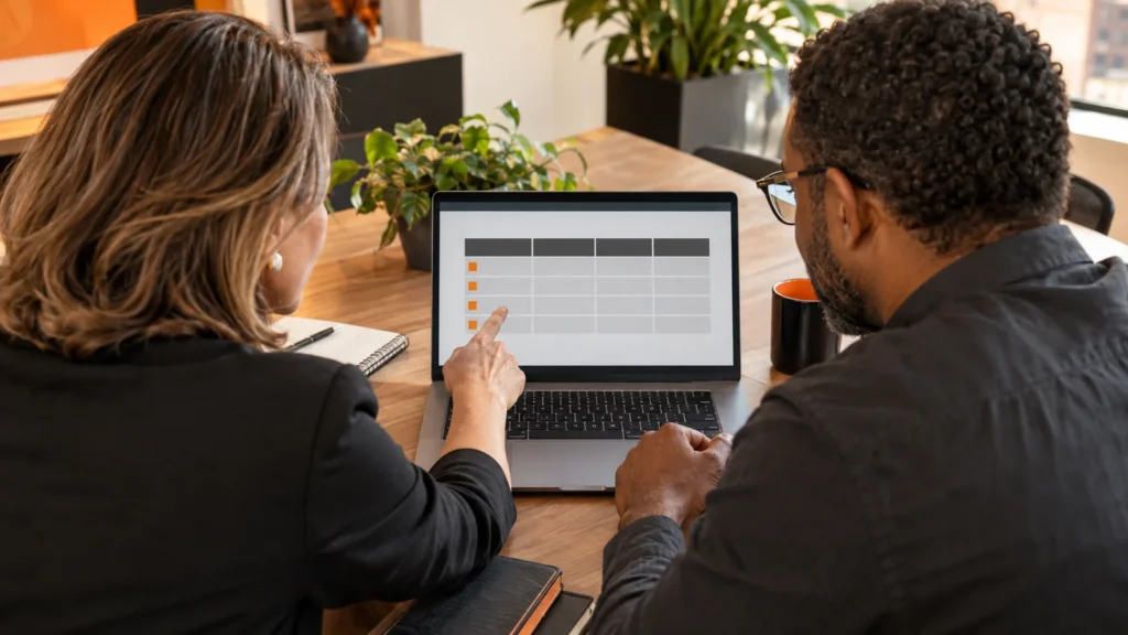 Gestora e analista observam juntos uma tabela na tela de um notebook sobre uma mesa de madeira.