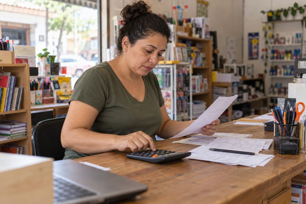 Mulher confere documentos e usa uma calculadora em uma mesa de madeira numa papelaria.